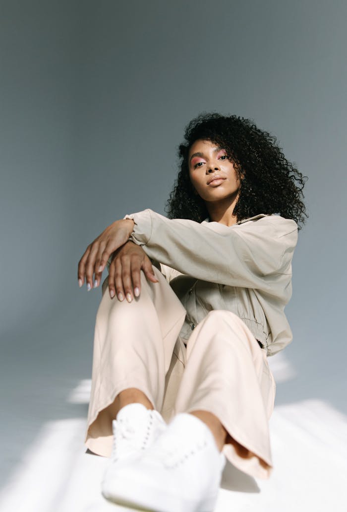 Stylish portrait of a woman with curly hair, sitting in soft natural light against a gray background.